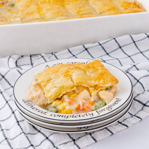 A slice of creamy chicken pot pie with a golden crust on a plate, showing visible vegetables, set on a stack of plates with text, beside a larger dish of pot pie on a checkered cloth.