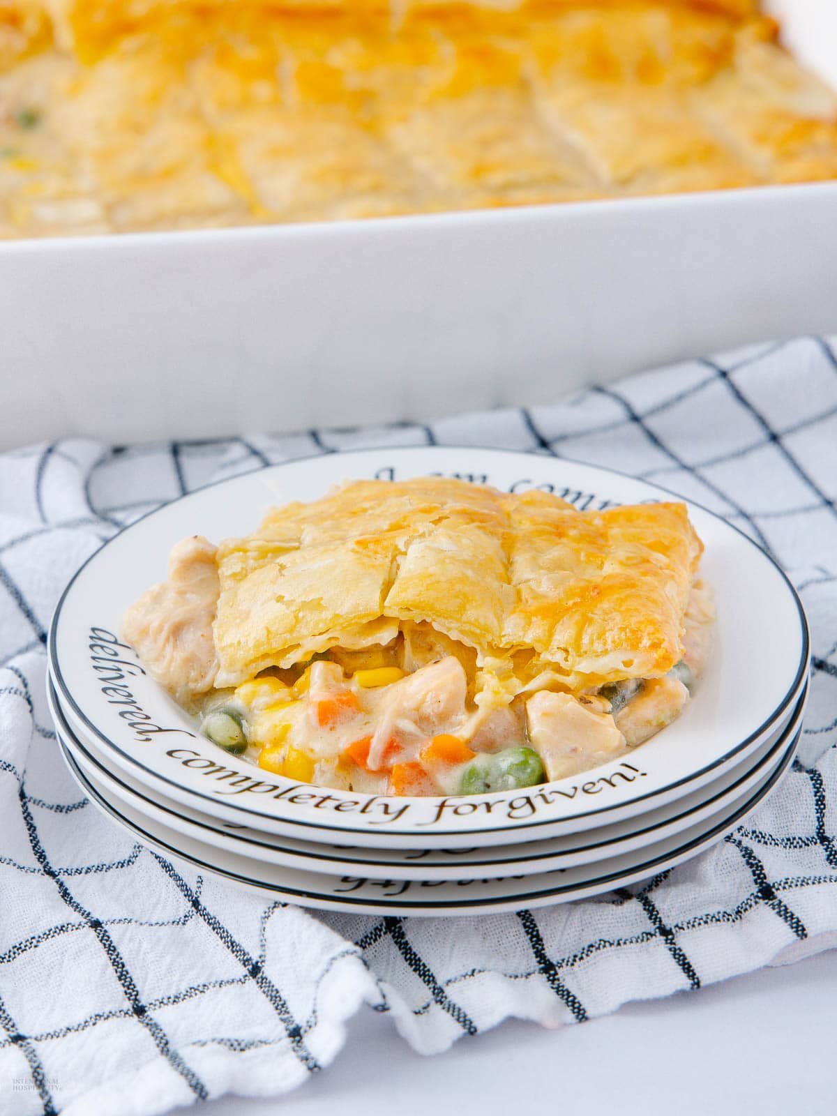 A slice of golden, flaky chicken pot pie rests on a stack of three white plates with black cursive text, set on a white and black checkered cloth. The pot pie is filled with chicken, peas, carrots, and sauce.