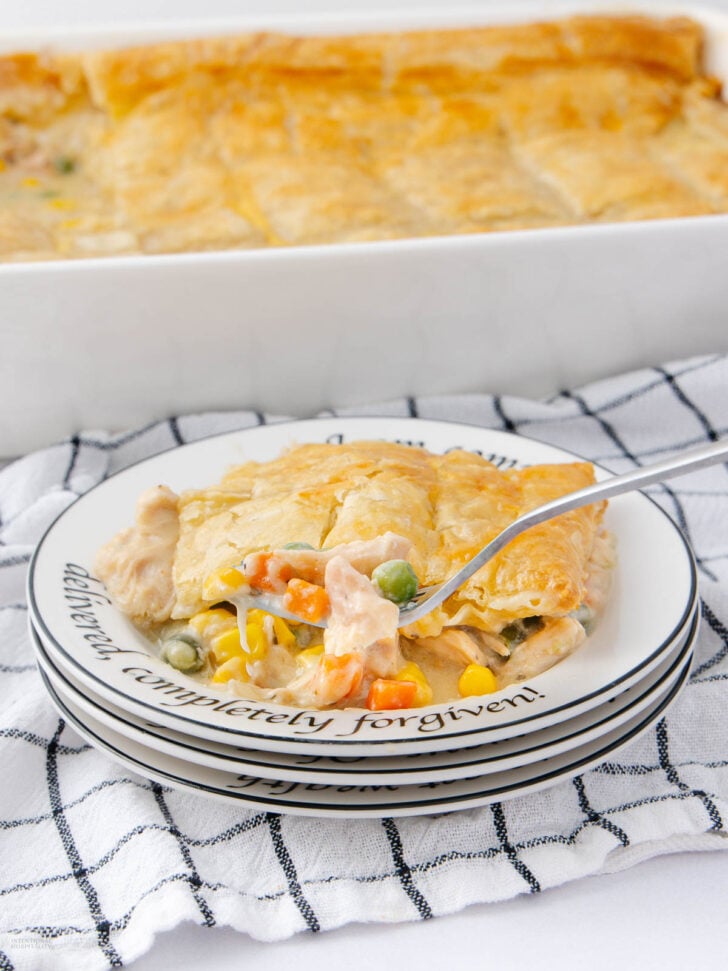A slice of creamy chicken pot pie with mixed vegetables on a plate, with a fork holding a bite. The plate sits on stacked dishes and a checkered towel, with a baking dish of the pot pie in the background.