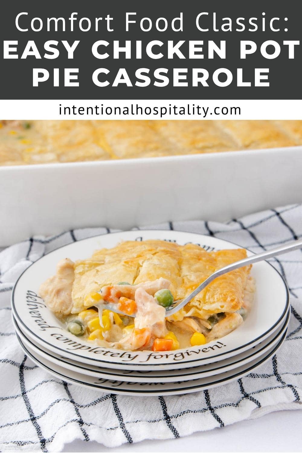 A slice of chicken pot pie casserole with vegetables is served on a plate with a fork. The plate rests on a stack of similar plates and a checkered cloth, with the rest of the casserole in the background.