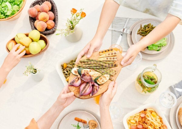 A table set for a meal with plates of pasta, salad, fresh fruits, and grilled vegetables on a wooden board. Multiple hands reach in to serve or share food, creating a warm, communal atmosphere.