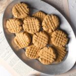 A silver oval plate holds several peanut butter cookies with crisscross fork marks, placed on an open book and a white surface.