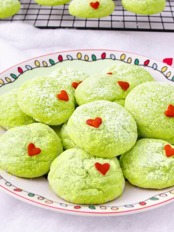 A plate of bright green cookies dusted with powdered sugar, each topped with a small red heart sprinkle. More cookies cool on a wire rack in the background. The plate has a colorful holiday light pattern around the rim.