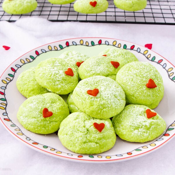 A plate of bright green cookies dusted with powdered sugar, each topped with a small red heart sprinkle. More cookies cool on a wire rack in the background. The plate has a colorful holiday light pattern around the rim.