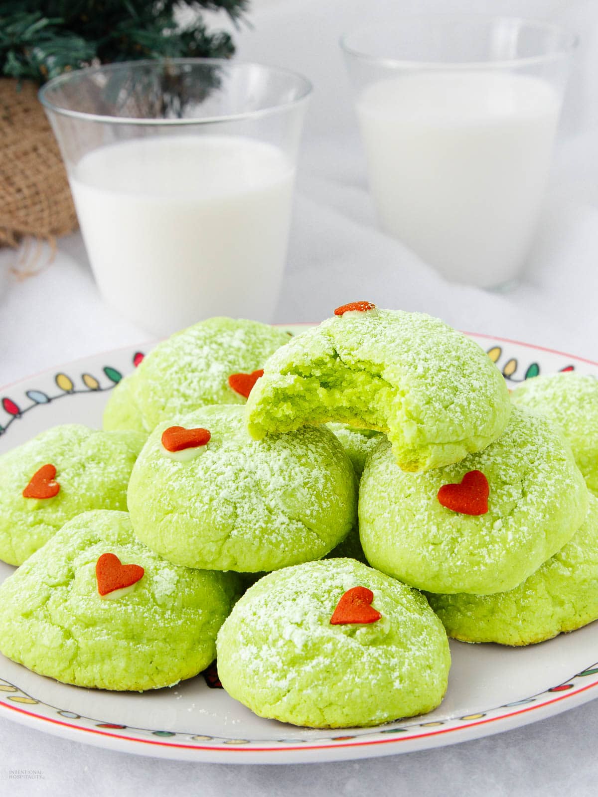A plate of green cookies dusted with powdered sugar and decorated with small red heart sprinkles, with two glasses of milk in the background. One cookie has a bite taken out of it.