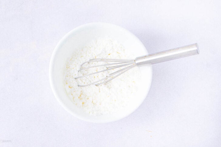 A metal whisk rests inside a white bowl filled with a white, powdery mixture, placed on a light-colored surface.