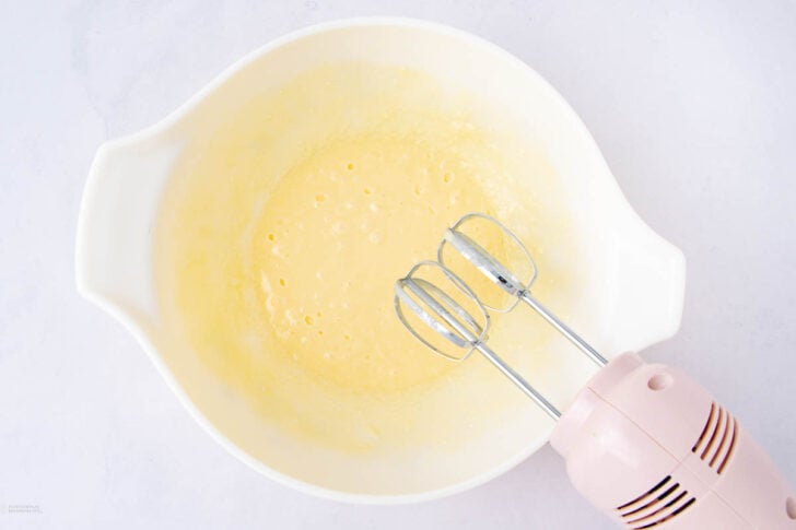 A white mixing bowl with pale yellow batter inside, being mixed with a pink electric hand mixer.