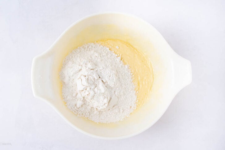A white mixing bowl containing a pile of flour on top of yellow cake batter, placed on a white surface.
