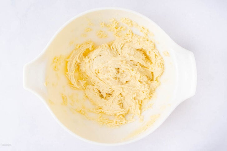A white mixing bowl containing creamed butter and sugar, with a light, fluffy texture, sits on a white surface.