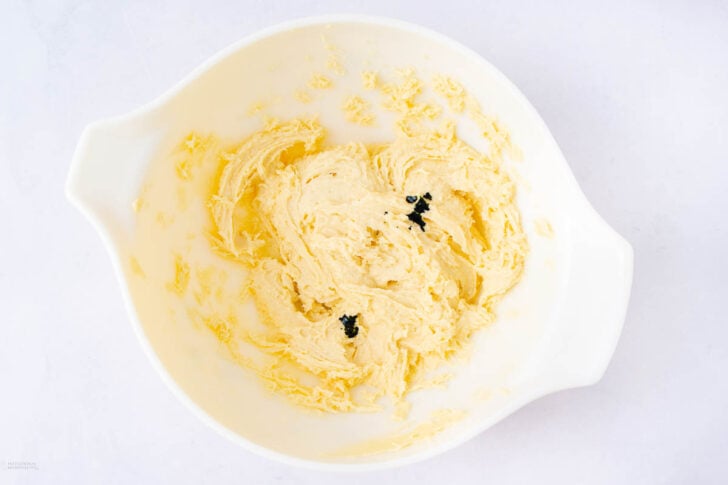 A white mixing bowl containing creamy, pale yellow batter with a few drops of dark liquid, possibly vanilla extract, on top. The bowl sits on a white surface.