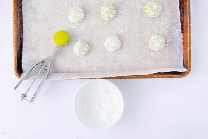 A baking tray lined with parchment paper holds seven round green dough balls coated in powdered sugar. An ice cream scoop with dough rests on the tray’s edge. A white bowl filled with powdered sugar sits below.