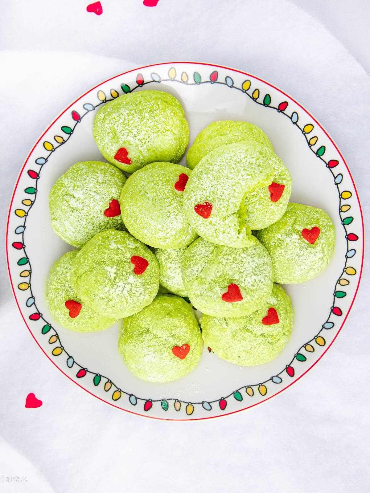 A plate of Grinch cake mix cookies dusted with powdered sugar, each topped with a small red heart-shaped sprinkle, arranged on a festive plate with colorful string light designs.
