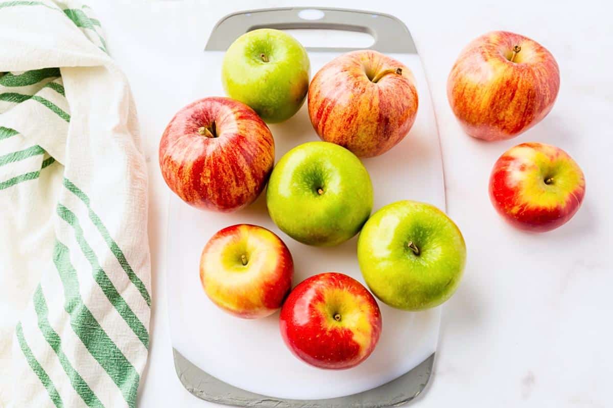 A white cutting board with five red apples and three green apples on it, next to a white towel with green stripes, placed on a white surface.