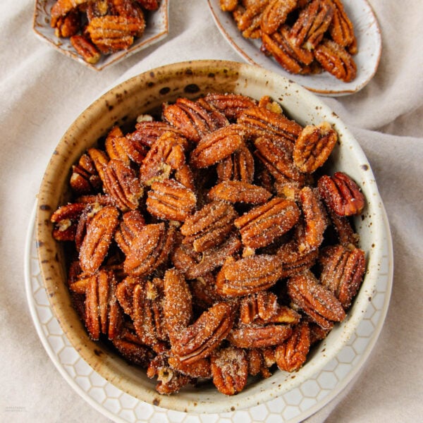 A bowl filled with candied pecans coated in sugar, with additional small plates of candied pecans in the background on a light-colored fabric surface.