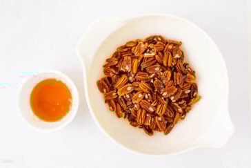 A white bowl filled with pecans sits next to a small white cup containing honey, all placed on a white surface.
