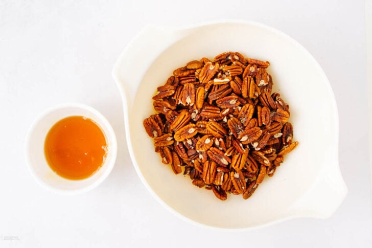 A white bowl filled with pecans sits next to a small white cup containing honey, all placed on a white surface.