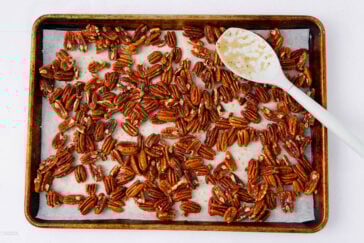 A baking sheet lined with parchment paper holds roasted pecans, some glistening with seasoning. A white mixing spoon rests on the sheet, coated with seasoning remnants. The background is plain white.