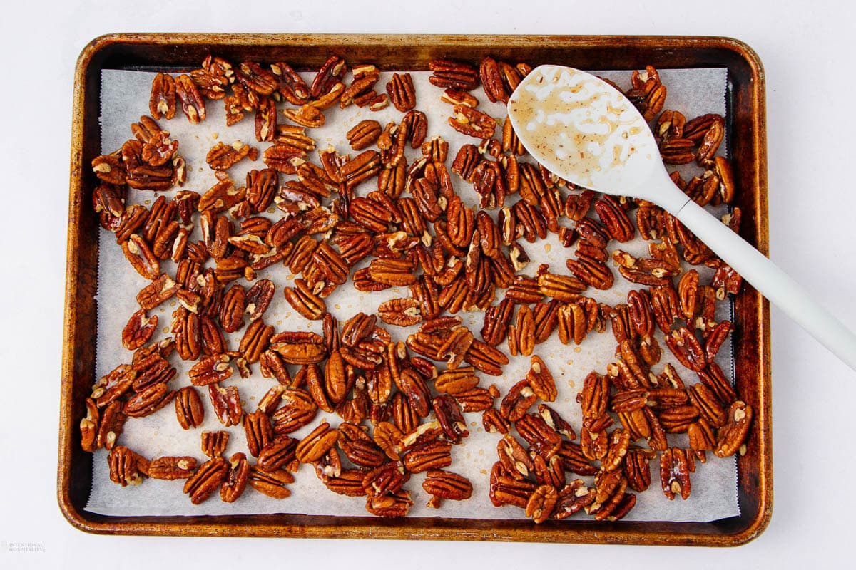 A baking sheet lined with parchment paper holds roasted pecans, some glistening with seasoning. A white mixing spoon rests on the sheet, coated with seasoning remnants. The background is plain white.