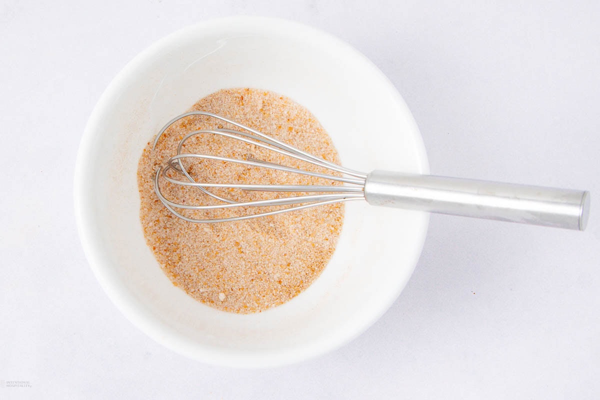A metal whisk rests in a white bowl containing a light brown seasoning or spice mixture on a white surface.