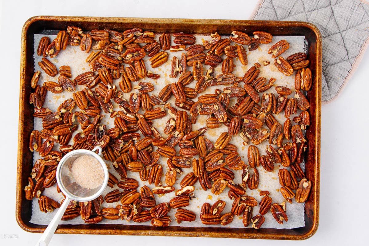 A baking sheet lined with parchment paper holds roasted pecans. A metal strainer filled with cinnamon sugar rests on the sheet, and an oven mitt is partially visible in the upper right corner.