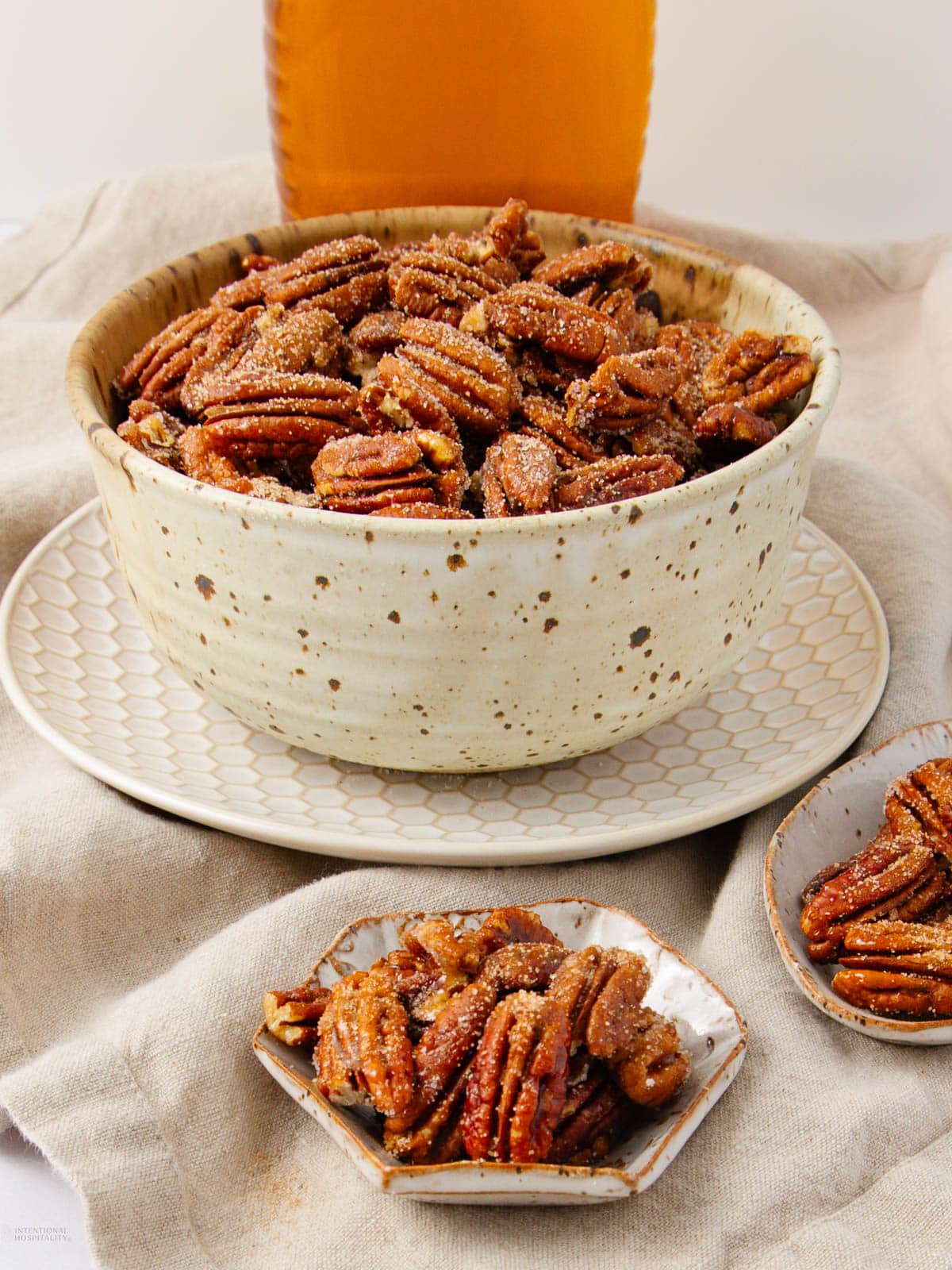 A rustic ceramic bowl filled with sugared pecans sits on a patterned plate, with extra pecans on small dishes nearby. A bottle of amber liquid is in the background, all atop a beige cloth.
