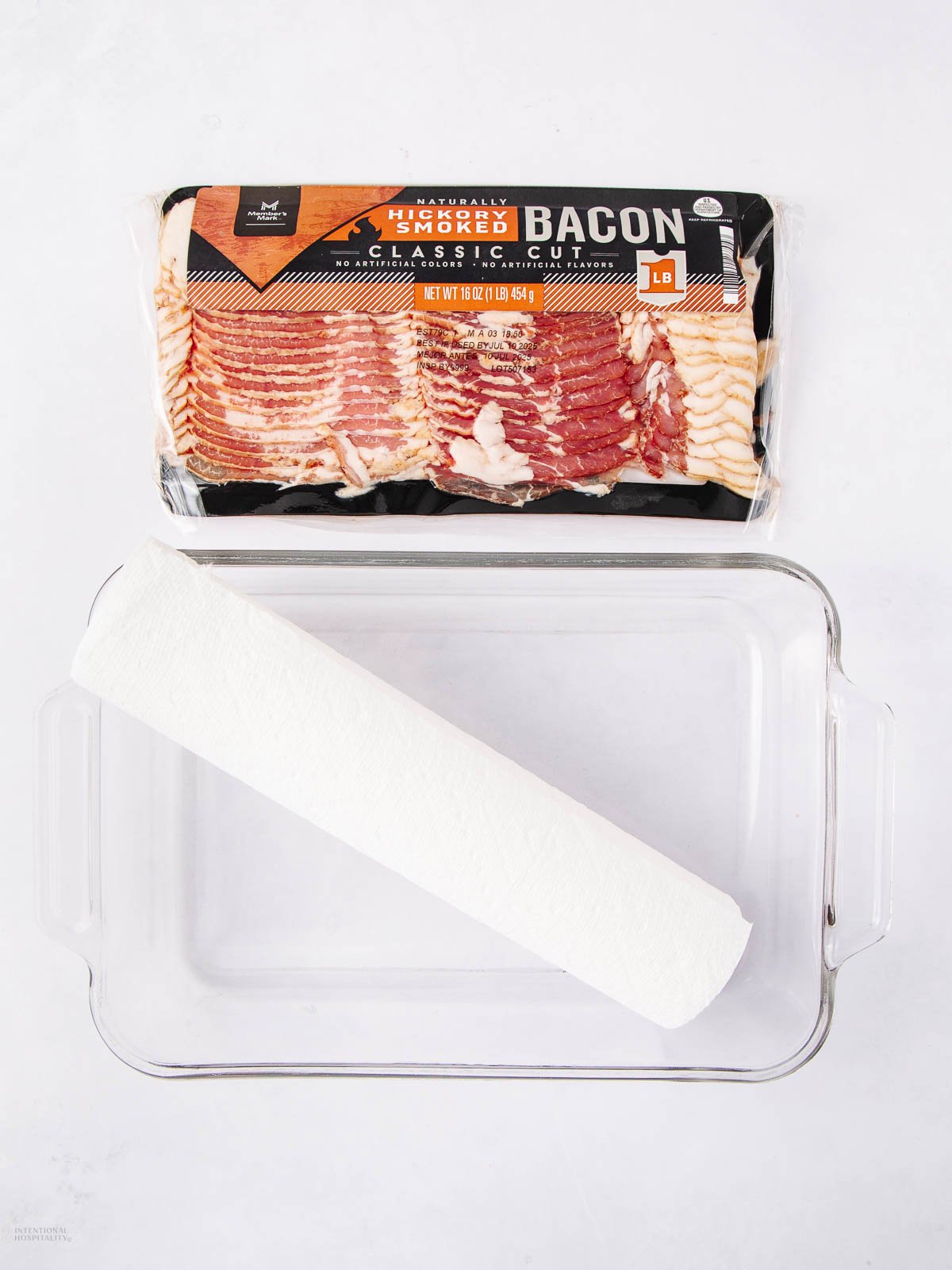 A package of classic cut smoked bacon, a clear glass baking dish, and a roll of white paper towels are arranged on a white surface.