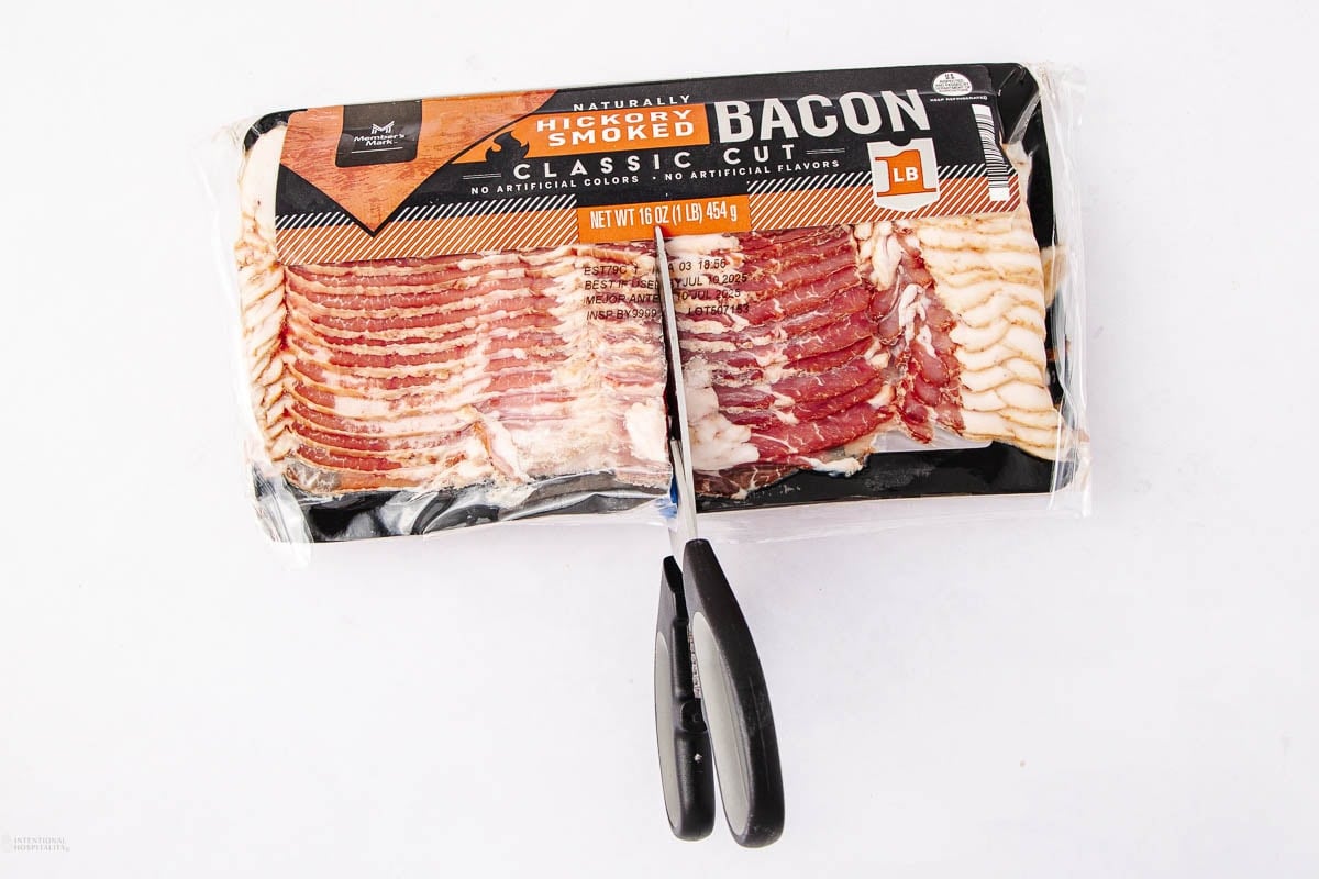 A pair of scissors is cutting through the middle of a sealed package of hickory smoked classic cut bacon on a white background.