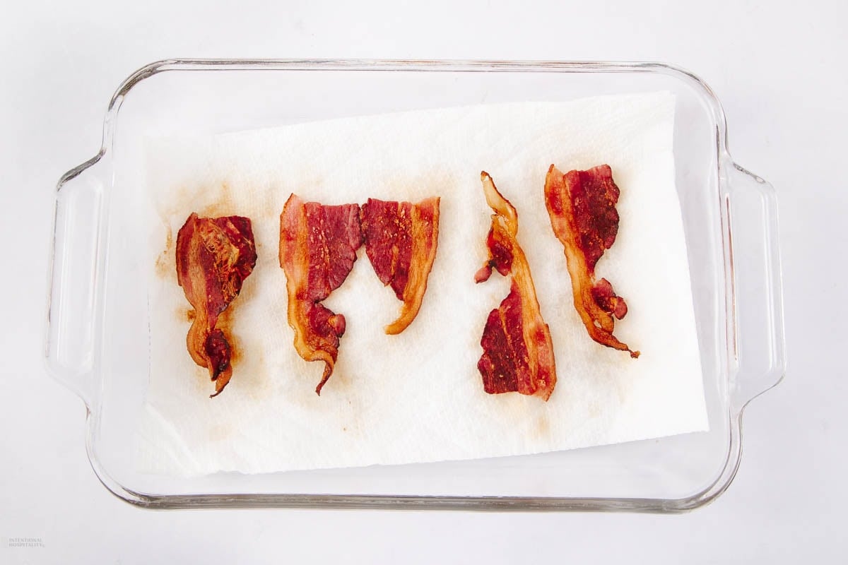 Five crispy cooked bacon strips rest on a white paper towel inside a clear rectangular glass baking dish, with some grease spots visible on the paper towel.