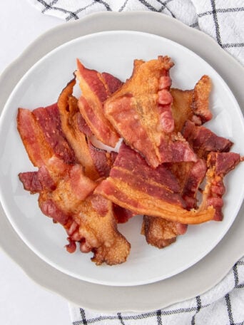 A white plate with crispy cooked bacon strips sits on a gray charger plate, next to a white and black checkered kitchen towel.