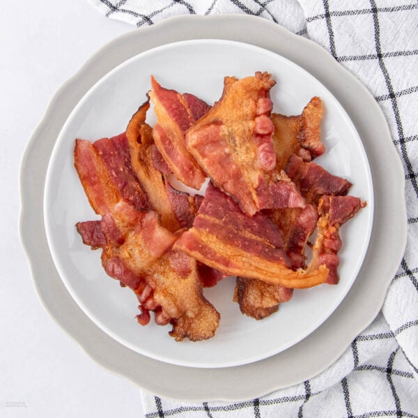 A white plate with crispy cooked bacon strips sits on a gray charger plate, next to a white and black checkered kitchen towel.