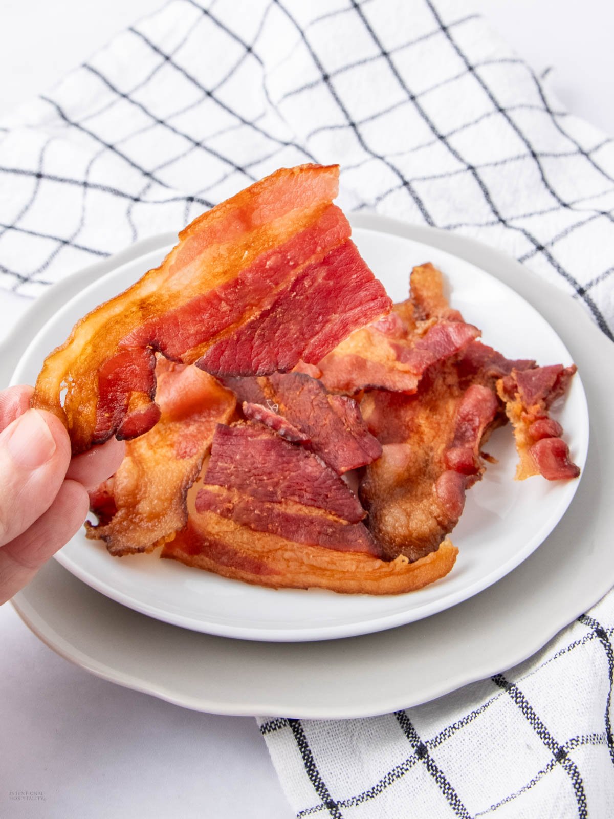 A hand holds a crispy, cooked strip of bacon above a small white plate filled with more bacon slices. The plate rests on a white cloth with a black checkered pattern.