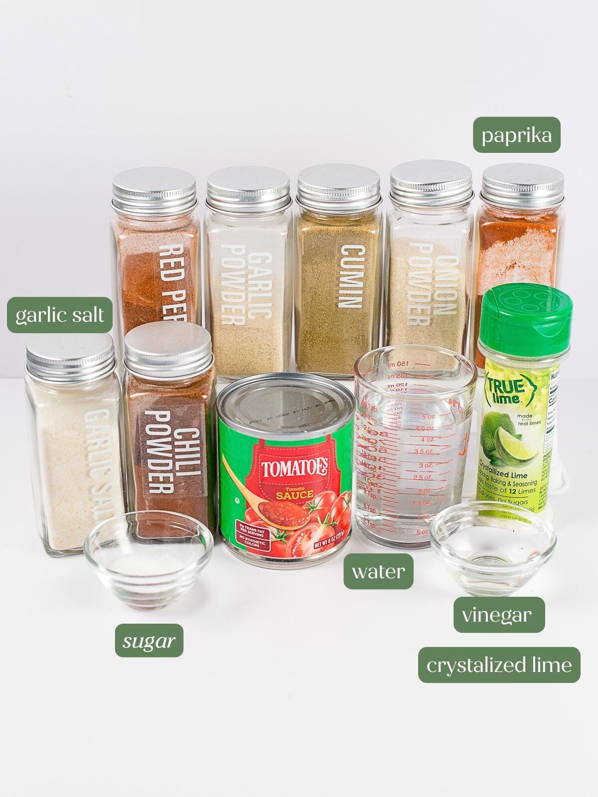 Assorted spice jars labeled red pepper, garlic powder, cumin, onion powder, paprika, garlic salt, chili powder, and sugar, plus tomato sauce, a measuring cup with water, a small bowl of vinegar, and crystallized lime.