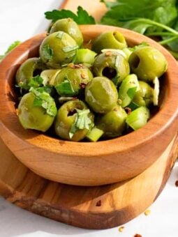 A wooden bowl filled with green olives garnished with chopped herbs and small pieces of garlic, placed on a wooden board with fresh parsley and garlic cloves in the background.