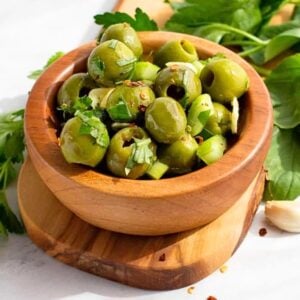 A wooden bowl filled with green olives garnished with chopped herbs and small pieces of garlic, placed on a wooden board with fresh parsley and garlic cloves in the background.