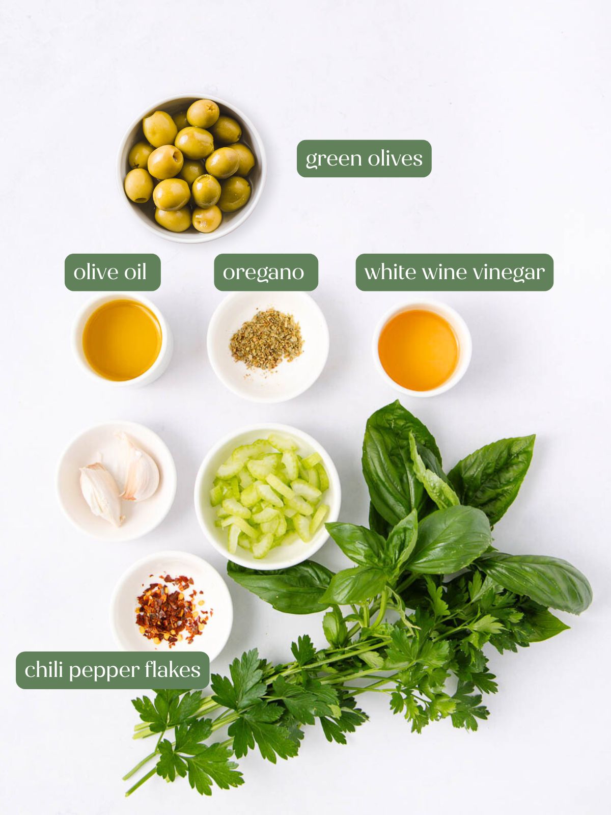A flat lay of ingredients on a white surface: green olives, olive oil, oregano, white wine vinegar, chili pepper flakes, garlic cloves, chopped celery, fresh basil, and fresh parsley, each labeled with text.