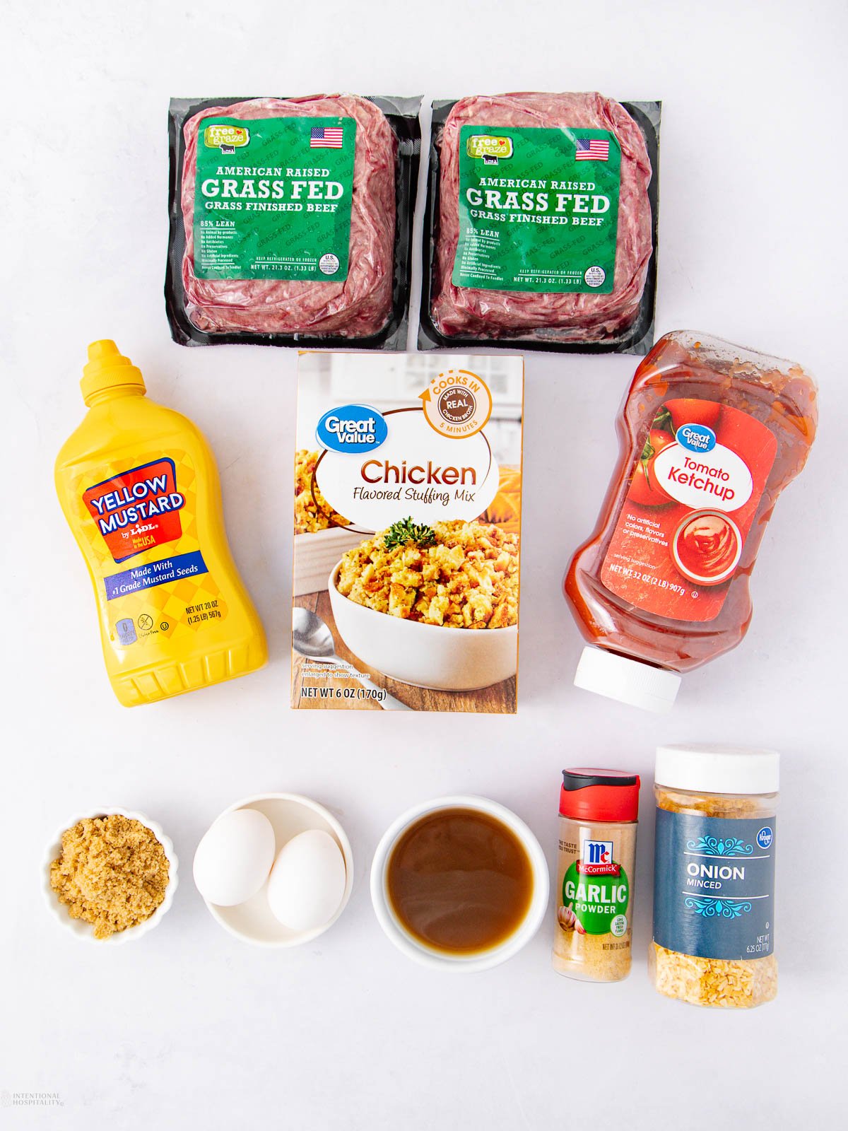 Ingredients for a meatloaf on a white surface: two packs of ground beef, chicken stuffing mix, ketchup, yellow mustard, brown sugar, two eggs, minced garlic, dried onion, and a bowl of brown liquid.