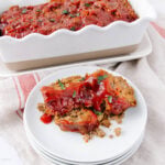 Two slices of meatloaf topped with ketchup glaze are served on a stacked white plate, with the rest of the meatloaf in a white loaf pan in the background, set on a light-colored cloth.