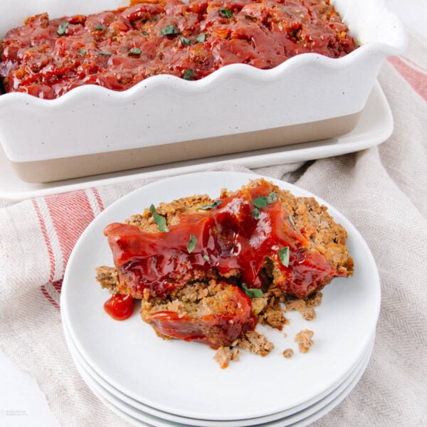 Two slices of meatloaf topped with ketchup glaze are served on a stacked white plate, with the rest of the meatloaf in a white loaf pan in the background, set on a light-colored cloth.