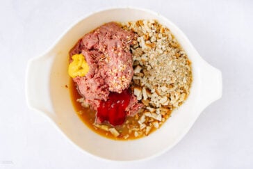 A white bowl containing ground meat, breadcrumbs, crushed crackers, diced onions, ketchup, mustard, and other seasonings, ready to be mixed for a recipe.