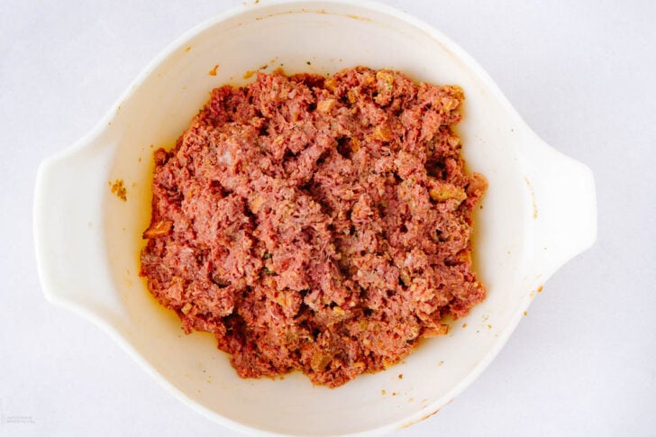 Raw ground meat mixture with seasonings in a white mixing bowl, ready to be cooked or shaped, placed on a white surface.