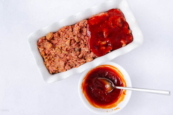 Overhead view of a rectangular white dish with raw meatloaf, half covered in red sauce. A small white bowl of red sauce with a spoon rests beside it on a white surface.