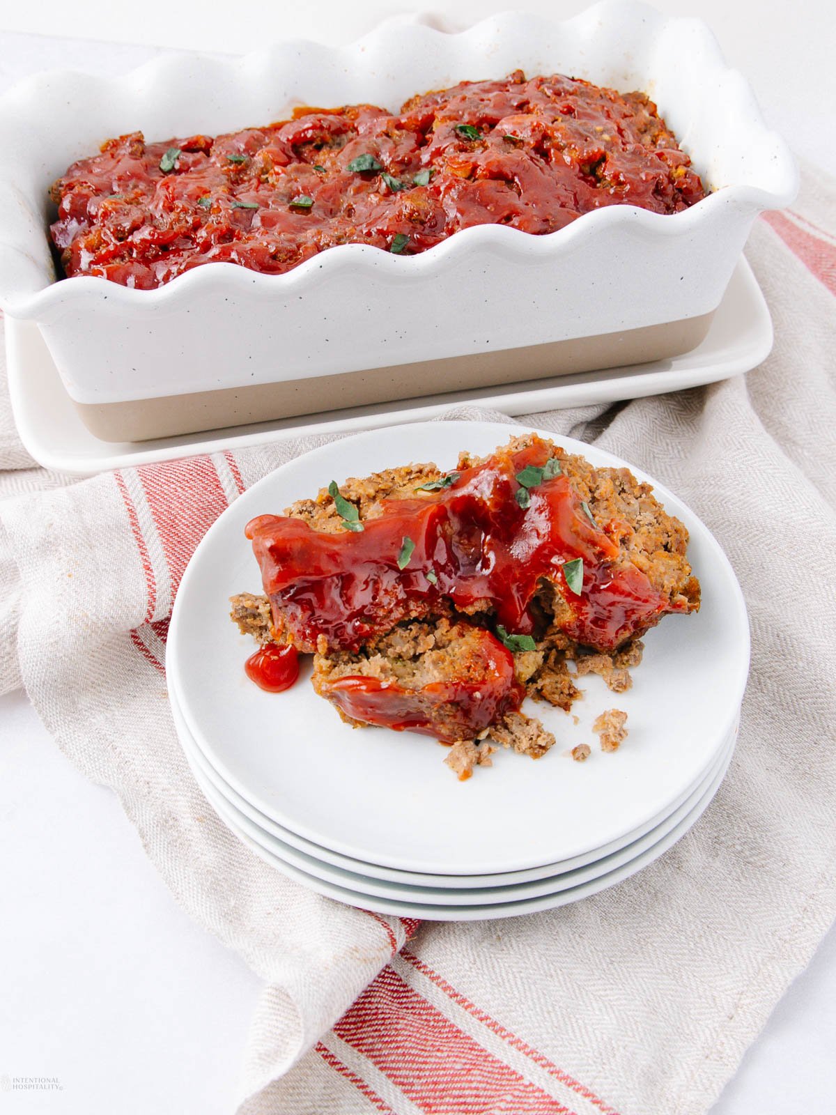 Sliced meatloaf topped with ketchup glaze on a white plate, with the remaining meatloaf in a white loaf pan behind it, set on a red-striped kitchen towel.