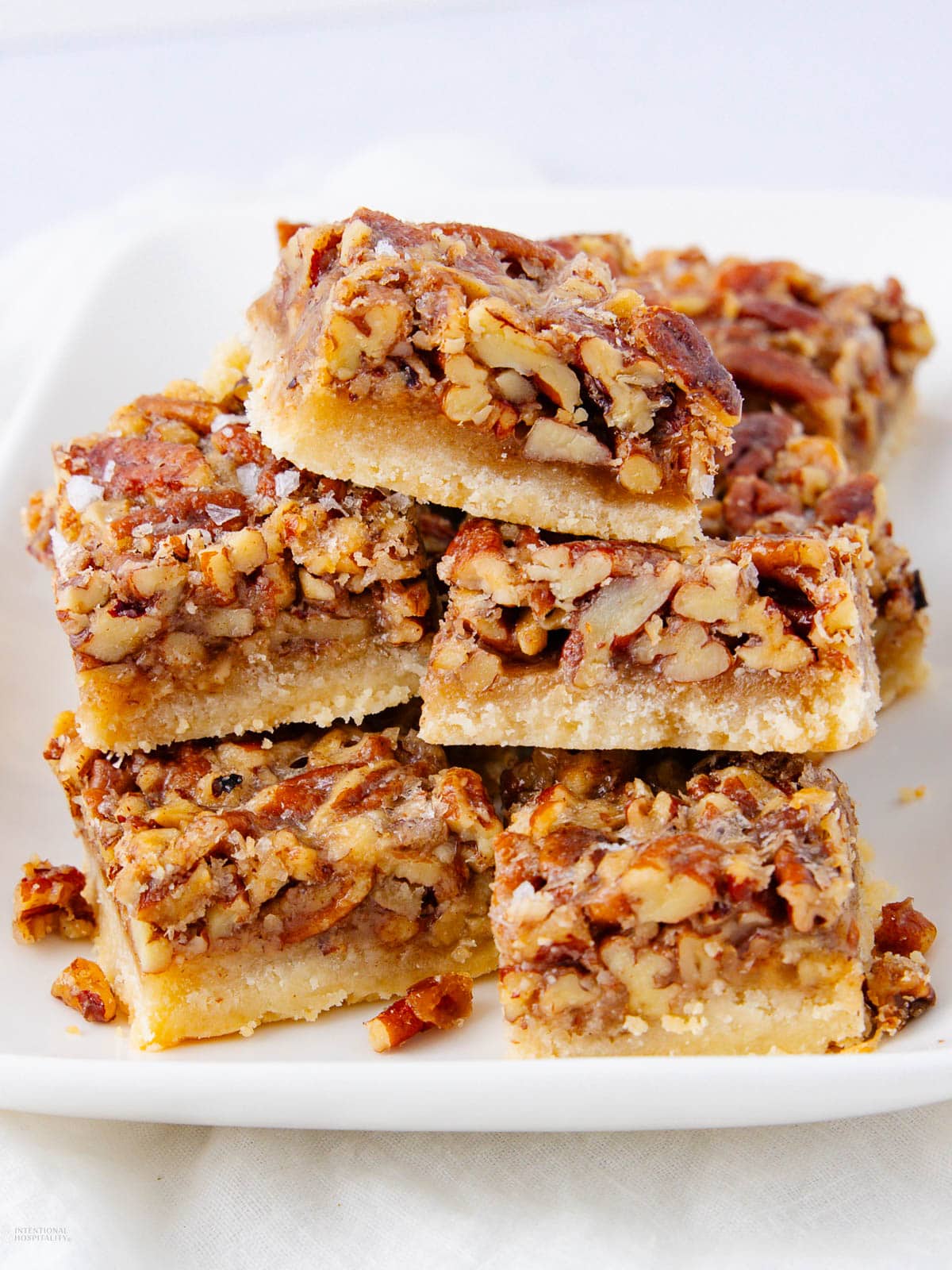 A plate stacked with pecan pie bars, featuring a golden shortbread crust topped with a thick layer of chopped pecans and caramelized filling. The bars are cut into squares and arranged neatly on a white plate.
