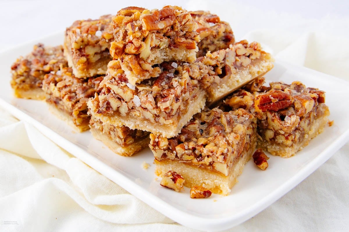 A white plate with stacked pecan bars featuring a crumbly, golden crust and a dense topping of chopped pecans and caramelized filling, set on a white cloth background.