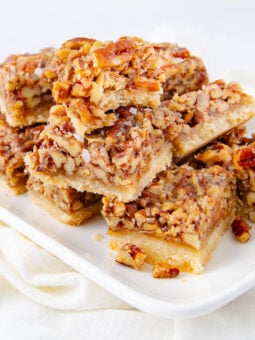 A white plate with several stacked pecan bars, featuring a golden crust and a generous layer of chopped pecans in a caramel topping. The plate rests on a light-colored cloth.
