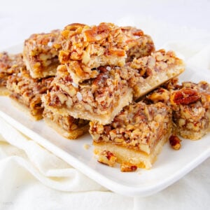 A white plate with several stacked pecan bars, featuring a golden crust and a generous layer of chopped pecans in a caramel topping. The plate rests on a light-colored cloth.