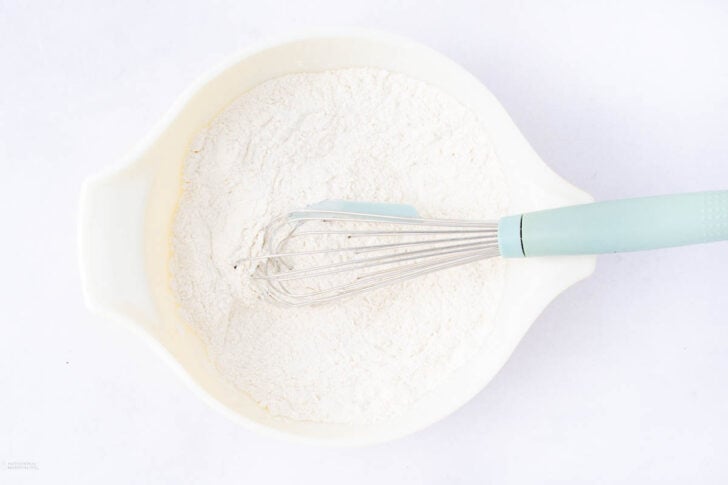 A white mixing bowl filled with flour, with a light blue-handled whisk resting inside on a white background.