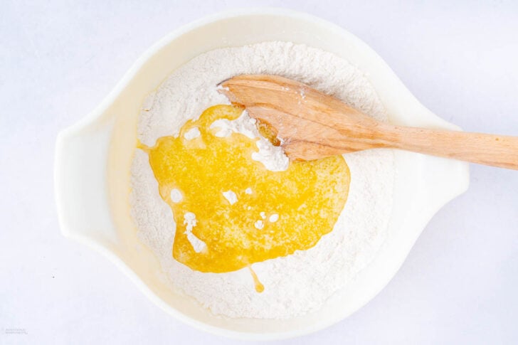 A white bowl containing flour and melted butter, with a wooden spoon resting in the mixture, viewed from above on a white background.