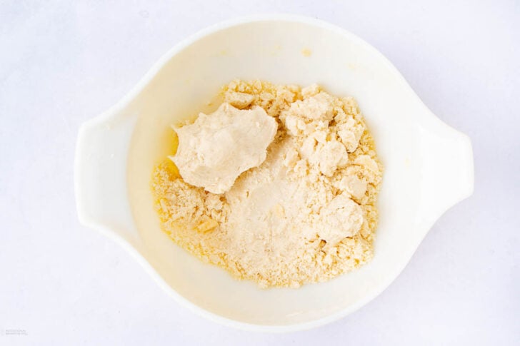 A white mixing bowl containing crumbly dough, with a portion forming a soft, smooth ball and the rest remaining loose and powdery. The bowl is set on a white surface.