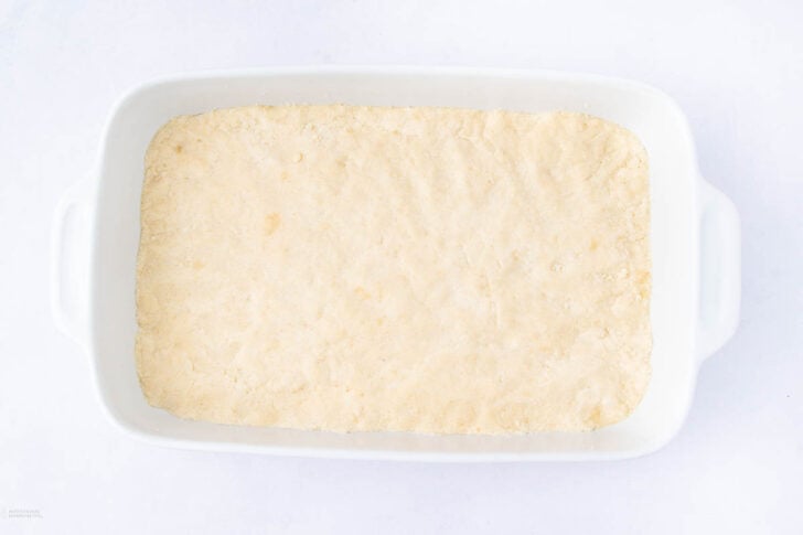 A rectangular white baking dish filled with an even layer of unbaked dough, pressed flat across the bottom, on a white background.